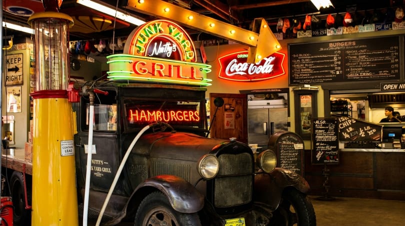 Vintage gas pump and antique truck with neon signs for "Nutty's Junkyard Grill" and "Hamburgers" inside a diner.