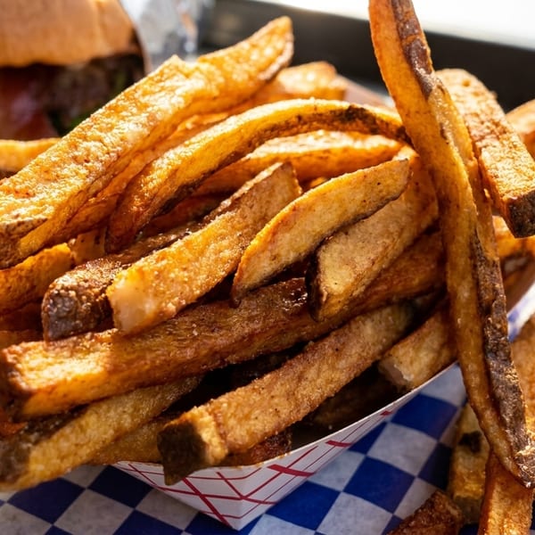 Crispy golden french fries in a checkered paper tray, with a burger blurred in the background.