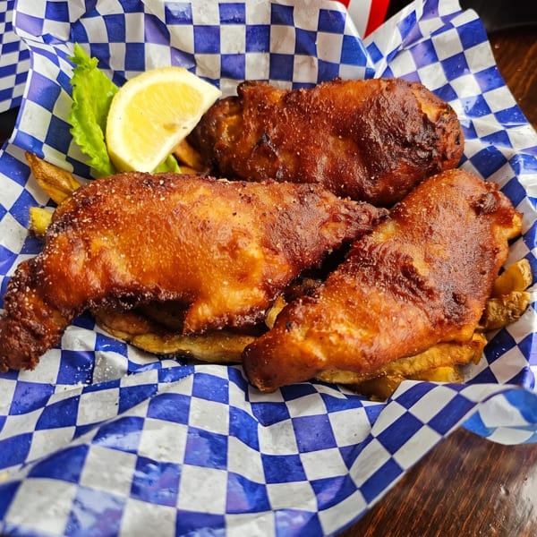 Three pieces of golden-brown fried fish with fries, lemon, and lettuce in a blue checkered basket.