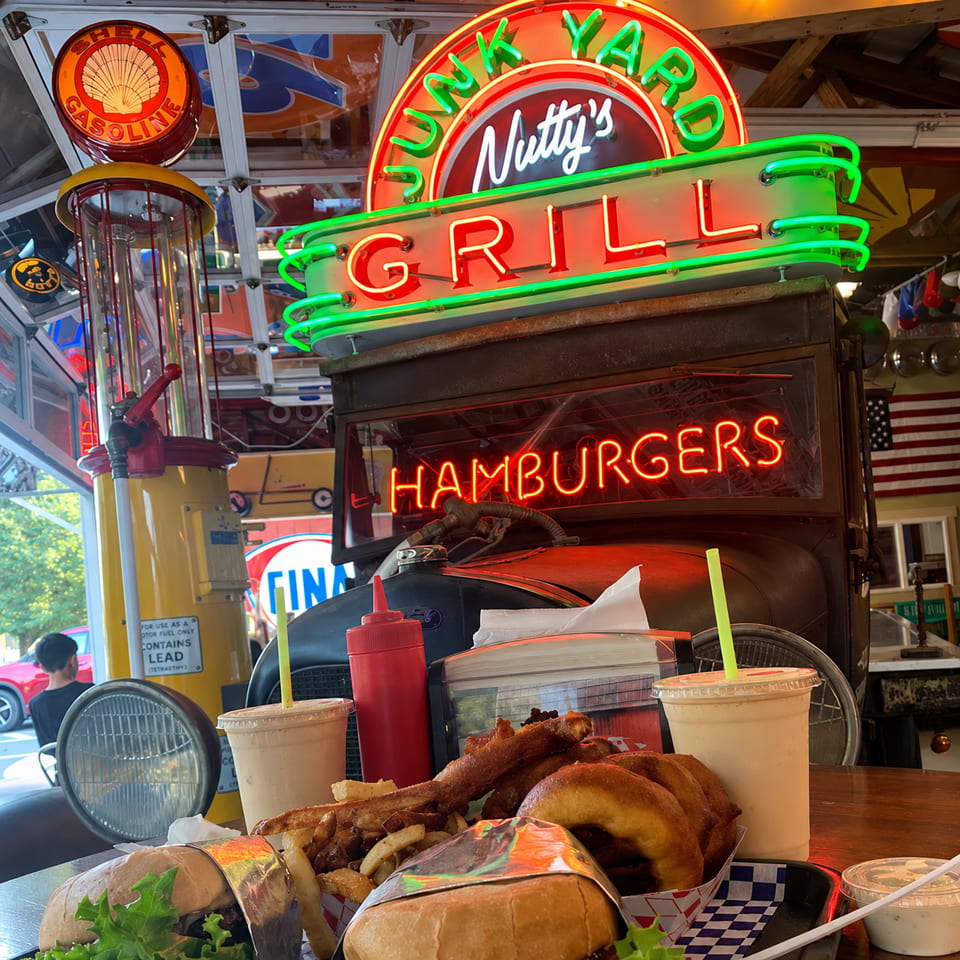 Nutty's Junkyard Grill neon sign above a table with burgers, fries, and milkshakes.