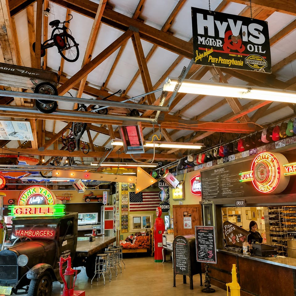 A rustic restaurant interior with vintage decor, neon signs, an old truck, and a counter with a server.