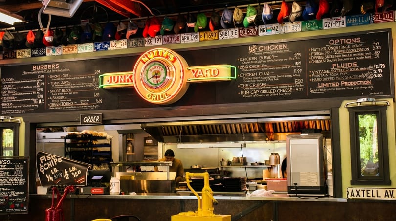 A brightly lit diner counter with a neon sign for "Nutty's Junkyard Grill" above a menu board.