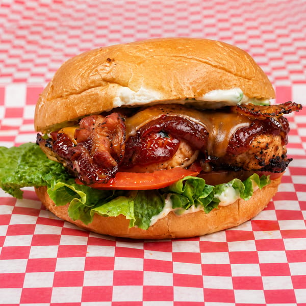 Grilled chicken sandwich with melted cheese, bacon, BBQ sauce, lettuce, and tomato on a bun.