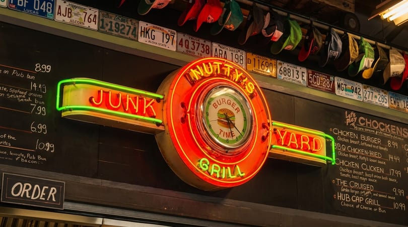 A vibrant neon sign for "Nutty's Junk Yard Grill" with a "Burger Time" clock, above a menu board.